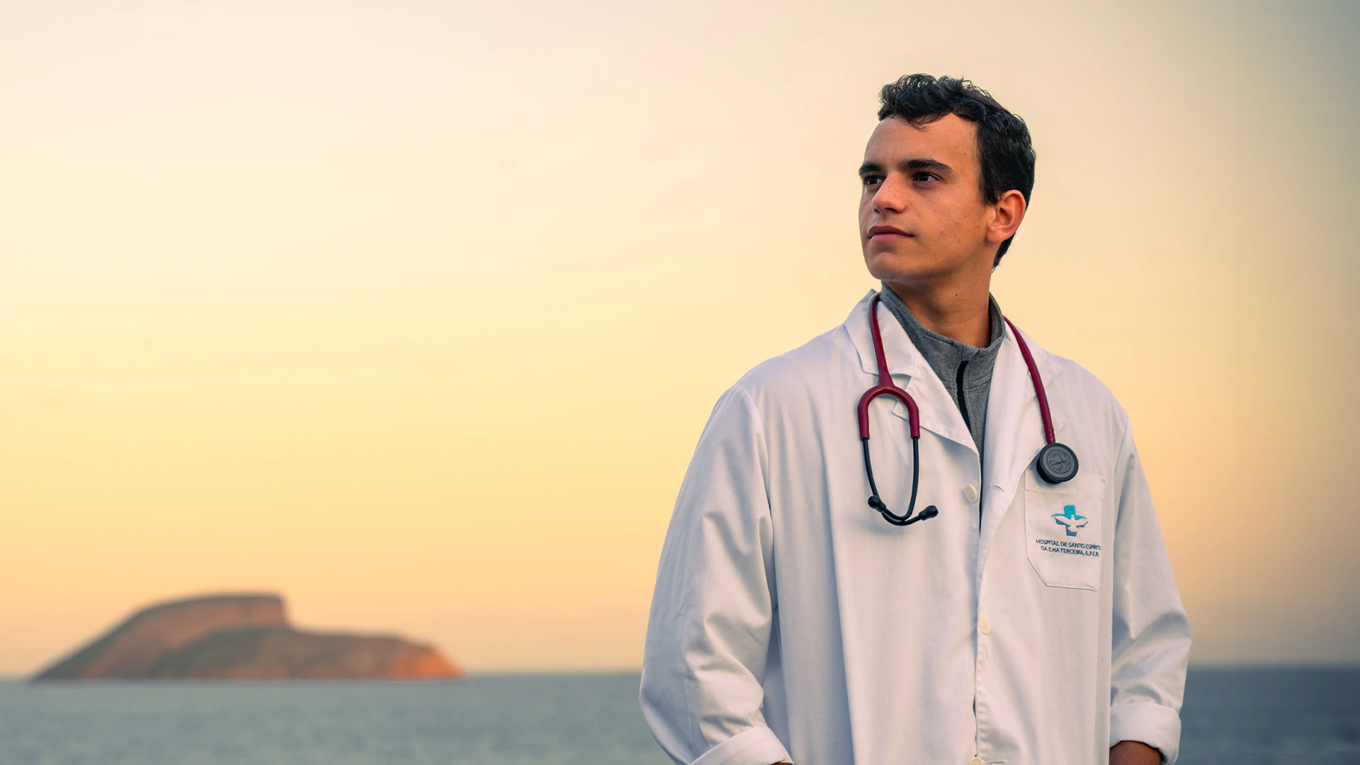 Médico com bata e estetoscópio, com o mar e uma ilha dos Açores ao fundo